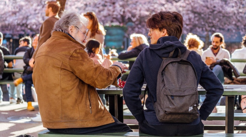 FOTO — zwei Menschen an einem Gemeinschaftstisch (Schach-Szene), draußen im warmen Sonnenlicht, mit Menschen im Hintergrund. Natürlich und unvermittelt, Motive leicht links der Mitte. 16:9.